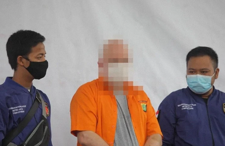 Mies oranssissa vankilahaalarissa kahden indonesialaisen poliisin välissä, kasvot peitettyinä